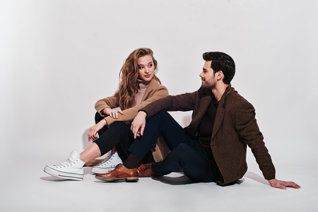Always in style. Attractive and well-dressed couple posing in studio. They are looking at each other, while sitting on the floor. Isolated over grey backgroundの写真素材