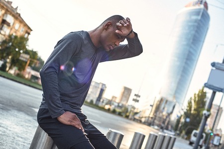 Need a break. Tired african athlete touching forehead and resting during urban outdoor workoutの写真素材
