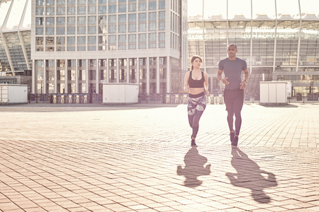 Positive and sporty mixed race couple: handsome african man and brunette girl in sportswear running together on a sunny warm day.の写真素材