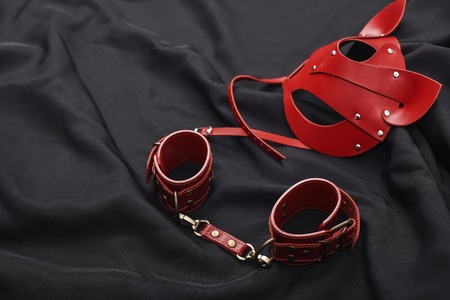 The Pleasures of BDSM. Top view of red bdsm leather set (handcuffs and mask) on a black silk