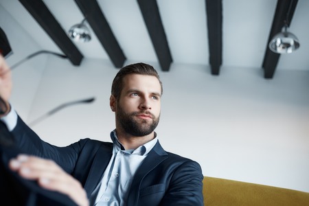 Close-up image of elegant and young businessman in suit who is holding glasses and looking aside...
