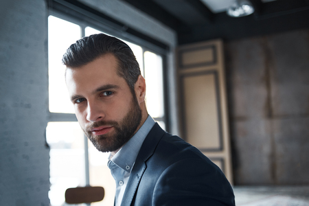 Being successful. Close-up portrait of sexy bearded young businessman who is looking at camera while standing in officeの写真素材