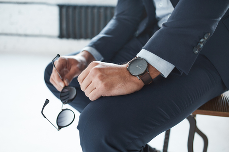Close-up image of a stylish businessman who is sitting on the chair with branded watch on his hand and holding glassesの写真素材