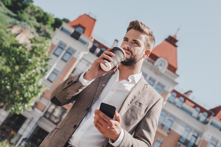 Coffee break on a go. Low angle view of confident brown-haired man holding coffee cup and using his smartphone while walking outdoorsの写真素材