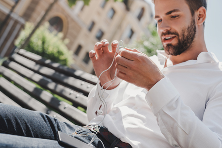 Sometimes the most productive thing you can do is relax. Handsome man holds his earphones while sitting on a bench in the parkの写真素材