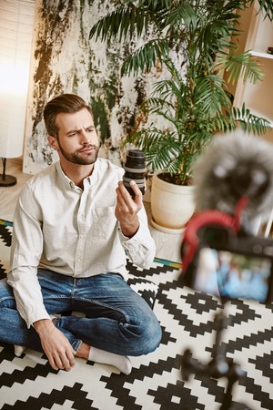 Technology blogger. Vertical portrait of young bearded blogger holding camera lens while recording new episode for his vlog.の写真素材