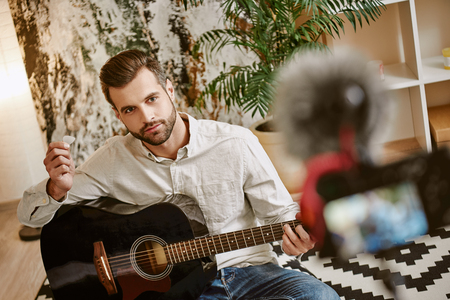 Learning guitar. Close up of attractive male music blogger sitting on the floor and showing how to play guitar with camera on foregroundの写真素材