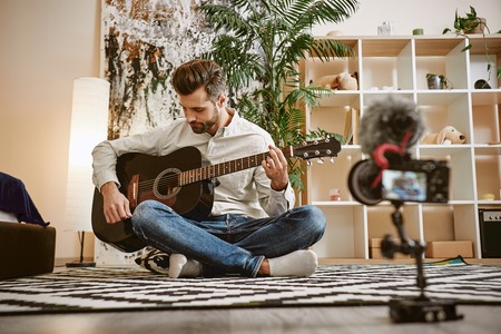 My inspiration. Bearded male music blogger sitting on the floor and holding the guitar, while recording new video for his vlogの写真素材
