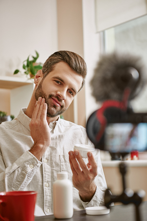 Soft skin. Vertical photo of positive male beauty blogger applying cosmetic cream on his face while live streaming.の写真素材