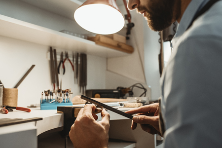 Focused on a process. Close up of a young male jeweler working and shaping an unfinished ring with a tool in workshop.の写真素材