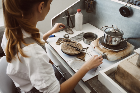 Thinking about new product. Side view of young female jeweler sitting at her jewelry making workshop and thinking which tools to useの写真素材