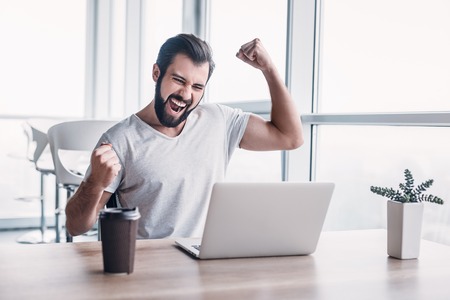 Handsome dark-haired businessman celebrating success with arms raised while looking at his laptop screen.の写真素材