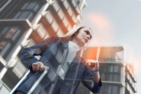 Trying to be in time. Beautiful young buiness woman in suit pulling luggage and checking the time while standing against of modern office buildings. Time management. Time concept. Business tripの写真素材