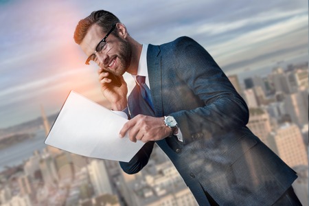Contract on good conditions. Cheerful young businessman talking by phone and looking at documents while standing against beautiful cityscape background. Partnership. Business concept. Successの写真素材