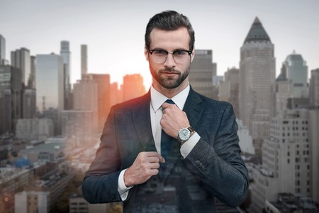 Perfect look! Confident business expert in glasses adjusting necktie while standing outdoors with cityscape on the background. Business look. Success conceptの写真素材