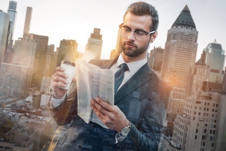 Reading news. Confident young businessman in suit reading newspaper and holding cup of coffee while standing outdoorsの写真素材