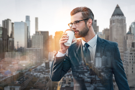 Coffee break. Stylish businessman drinking hot coffee and thinking about business while standing outdoors with cityscape on the backgroundの写真素材