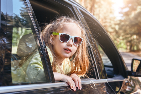 Cute cheerful little caucasian girl sitting inside the car, looking out the window. Family road tripの写真素材