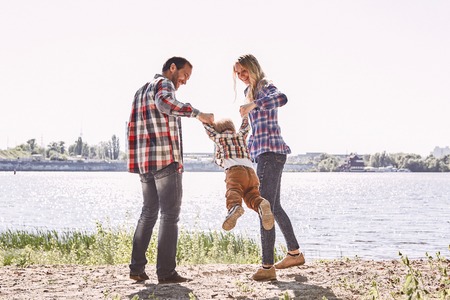 Family is not an important thing. Its everything. Parents standing with their kid, holding his hands and lifting him up. Summer landscape. Back viewの写真素材
