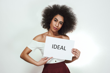 Studio portrait of confident young afro american woman holding a paper which is saying "Ideal" while standing against grey backgroundの写真素材