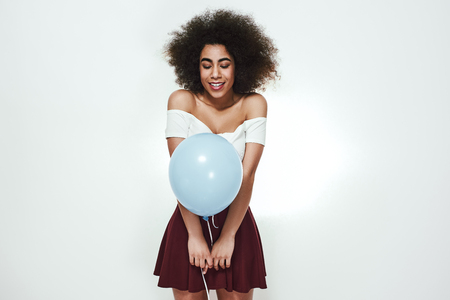 Cute and excited afro american woman with curly hair holding blue balloon with opened mouth. Studio shot. Birthday conceptの写真素材