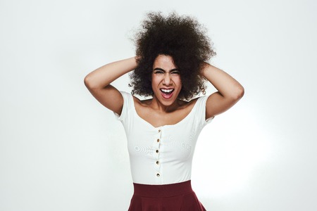 Playful and excited afro american woman touching her curly hair, looking at camera and screaming while standing against grey backgroundの写真素材