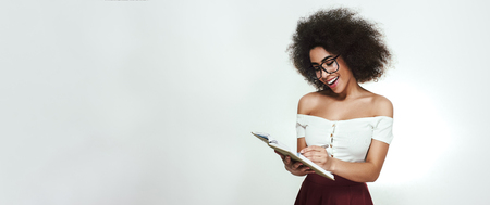 Cheerful and young african woman in eyewear writing something in her notebook while standing against grey backgroundの写真素材