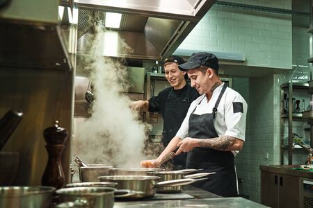 Side view of two cheerful and smiling cooks in uniform are preparing a food in a kitchen restaurantの写真素材