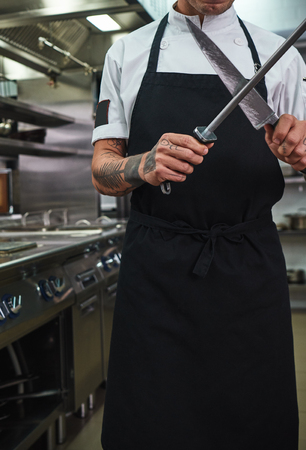 Ready to cook. Vertical photo of young man in apron with tattoos on his arms sharpening a knife in a restaurant kitchen.の写真素材