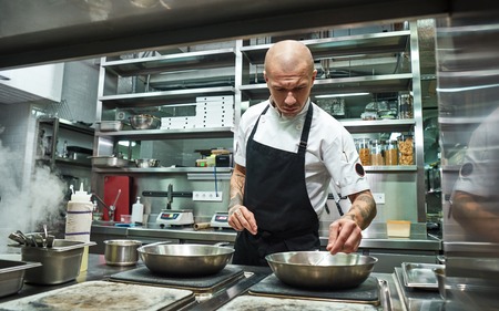 Confidence in everything. Serious male chef with several tattoos on his arms frying ingredients for his dish in a restaurant kitchen Confidence in everything. Serious male chef with several tattoos on his arms frying ingredients for his dish in a restaurant kitchen