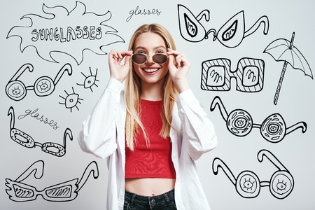 Beauty in sunglasses. Stylish and cheerful blonde woman in casual clothes wearing sunglasses while standing against grey background with hand drawn doodles on it.の写真素材