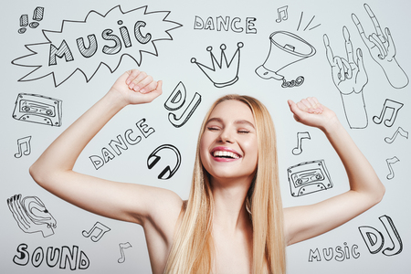 DJ party. Happy young blonde woman with naked shoulders smiling with raised hands while standing against grey background with music theme doodlesの写真素材