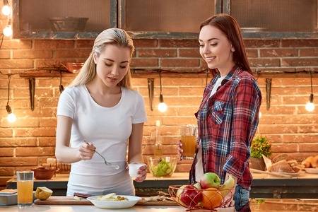 Portrait of two attractive women, having fun, while preparing fruit salad. They are fully involved in the process. Blonde girl in white T-shirt adds sour cream to chopped fruits in a bowl and her dark-haired friend in checkered shirt supervises the cooking processの写真素材