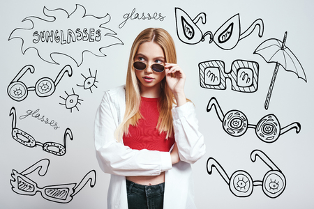 Fashion girl. Close up portrait of attractive asian woman adjusting her sunglasses and looking at camera while standing against grey background with hand drawn doodles on it. Fashion look. Beauty conceptの写真素材