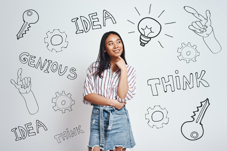 What if... Curious asian young woman in casual wear is smiling, touching her chin and and looking at drawn light bulb while standing against grey background with hand drawn doodles on it. Thinking concept. Idea conceptの写真素材