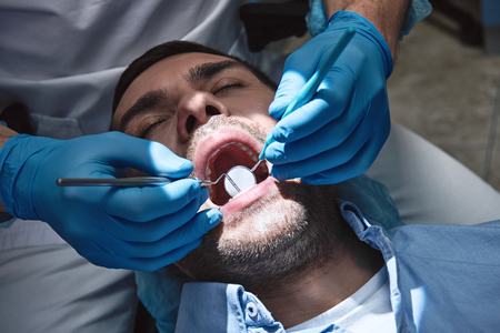 Bringing life to your smile. Dentist in latex gloves examining male patient, lying in dental chair with opened mouthの写真素材