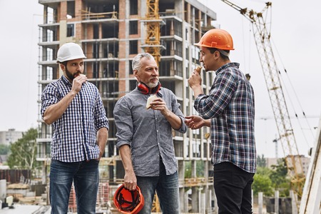 Lunch time. Group of builders in working uniform are eating sandwiches and talking while standing at construction site.の写真素材