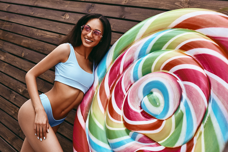 Salt in the air. Sand in my hair. African american model posing in bikini near big colorful lollipop isolated over wooden backgroundの写真素材