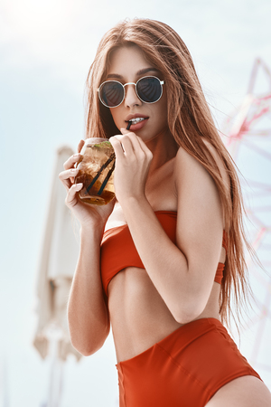 Take a dip in the pool, drink cocktail and stay cool. Portrait of slim fashion model in red swimsuit posing near the pool. Vertical shotの写真素材