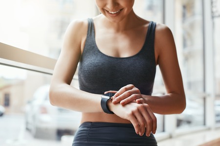 Checking all updates. Cropped photo of beautiful young woman looking at her sport bracelet while standing in front of window at gymの写真素材