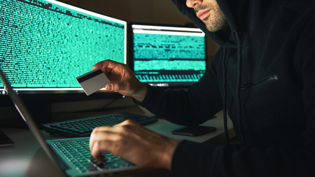 Stealing money. Side view of young bearded hacker in black hoodie using multiple computers and holding credit card while sitting in dark room. Binary code. Cyber attack. Cyber securityの写真素材