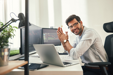 Young and successful. Happy bearded trader in formal wear and eyeglasses looking at camera and smiling while sitting in his modern office. Stock broker. Forex market. Trade conceptの写真素材