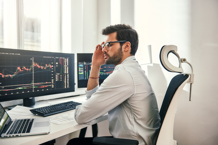 Full concentration. Young and smart businessman in formal wear is adjusting his eyeglasses and using laptop while while sitting in his office in front of computer screens with trading charts. Stock exchange. Trade concept. Investment conceptの写真素材
