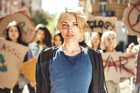 We believe in love. Young blonde woman with word love written on her face standing in front of female activists on the road during protest march.の写真素材