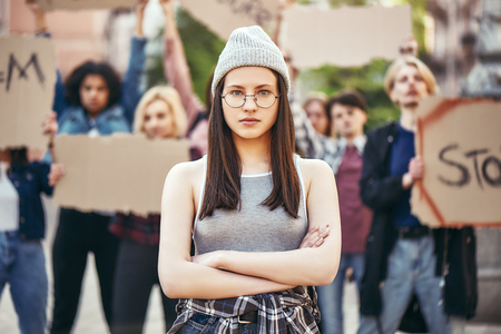 The future is female. Young woman in eyewear is standing on the road with crossed arms during a protest in front of female demonstrators holding signboards.の写真素材