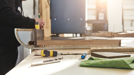 Close up of skilled carpenter in black uniform cutting a piece of wood in his woodwork workshop. Carpenter's tools lying on the table. Horizontal shot. Side view. Selective focusの写真素材