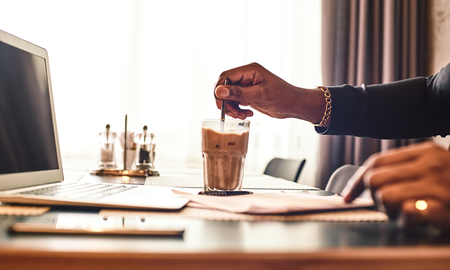 Close up of dark-skinned businessman in dark-blue jacket sitting at the table, indoors, drinking coffee. Young African entrepreneur puts sugar in his glass of coffee and mix it. Modern interiorの写真素材
