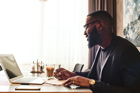 Portrait of dark-skinned businessman in dark-blue jacket and glasses sitting at the table, indoors, making notes, working with his laptop computer. Young African entrepreneur is coming up with new ideas. Modern interiorの写真素材