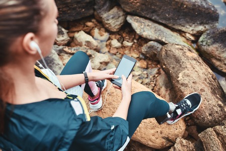 Disabled woman in sport wear with leg prosthesis holding phone in her hand and listening music while...