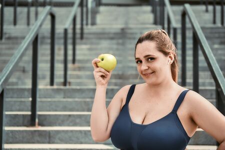 Healthy life. Positive plus size woman in sport clothing holding green apple and smiling while standing on stairs outsideの写真素材